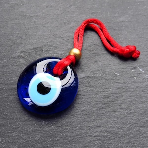 Può includere: Un ciondolo portafortuna di vetro blu con un design a occhio bianco e blu, appeso a uno stringa rossa con un perlina dorata.