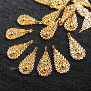 Può includere: Componenti per gioielli a forma di goccia dorata con intricata filigrana e una perla centrale. Questi ciondoli decorativi sono progettati per la creazione di gioielli e progetti di artigianato. I componenti sono disposti su una superficie scura.