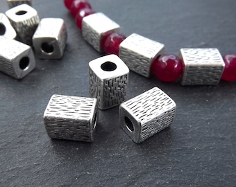 May include: Silver metal spacer beads with a textured finish. The beads have a square shape with a round hole in the center.