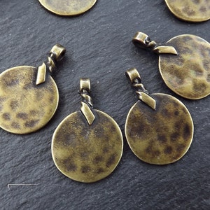 以下が含まれることがあります： ハンマー加工された質感と円形が特徴の、アンティークブロンズ色のペンダントがいくつかあります。各ペンダントの上部には、チェーンやコードに取り付けるためのねじれたループが付いています。ペンダントは暗い表面に散らばっています。