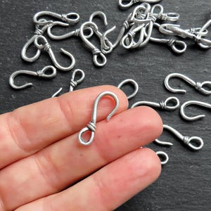 Op de afbeelding: Zilverkleurige metalen haken met een gedraaid draadontwerp. De haken zijn ongeveer 2,5 cm lang en hebben een lus aan de bovenkant om te bevestigen aan sieraden of andere knutselwerkjes.