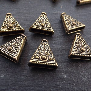 Könnte beinhalten: Ein Satz von acht antiken goldenen dreieckförmigen Metallperlen mit komplizierten Designs. Die Perlen haben eine erhabene Mitte und eine strukturierte Oberfläche.