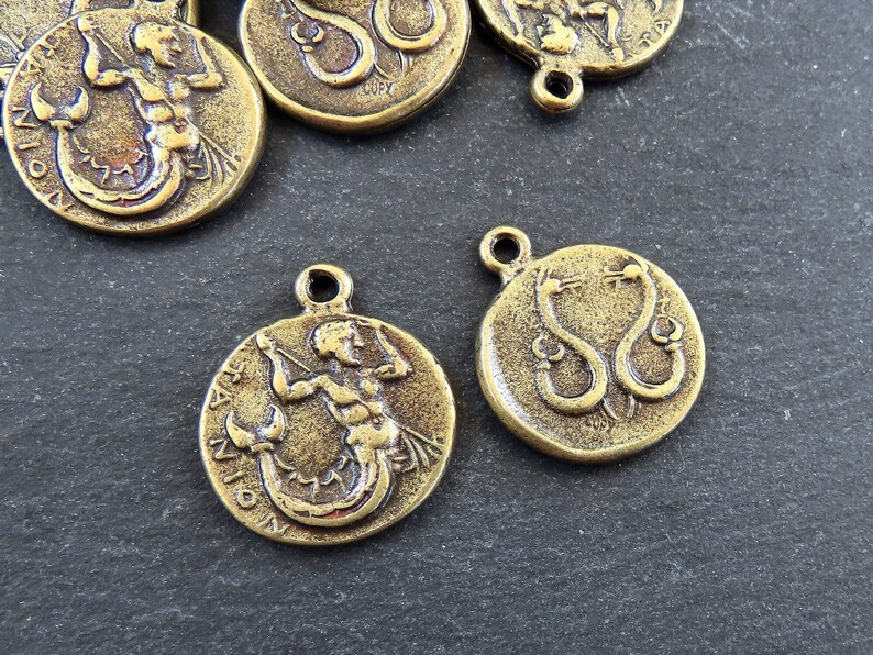 May include: Two gold-toned metal charms featuring zodiac signs. One charm depicts a mermaid with a fish tail and a bow and arrow, with the word "SAGITTARIUS" inscribed around the edge. The other charm depicts two snakes intertwined, with the word "SERPENS" inscribed around the edge.