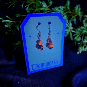 May include: A pair of dangle earrings with two orange and black speckled beads on each earring. The earrings are hanging on a blue card with the text "Domiworsk's" on it.
