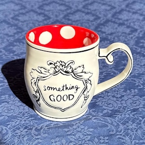 May include: A ceramic mug with a white exterior and a red interior featuring white polka dots. The front displays a black line-drawn crest containing the handwritten text something GOOD. The mug has a curved handle and a smooth, glossy finish.