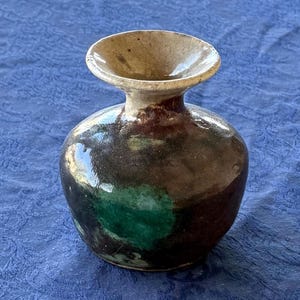 May include: A small, round ceramic vase with a wide, flared rim. The body of the vase features a glossy, dark brown glaze with a prominent, circular patch of vibrant green color on the front, and the neck transitions to a light beige tone.
