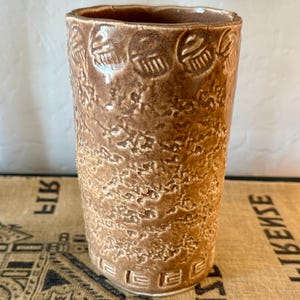 Może przedstawiać: Cylindryczny wazon ceramiczny z błyszczącym brązowym szkliwem. Zewnętrzna powierzchnia ma teksturowane, nakrapiane wykończenie, z pasem powtarzających się okrągłych odcisków przy górnej krawędzi oraz geometrycznym wzorem greckim u podstawy.