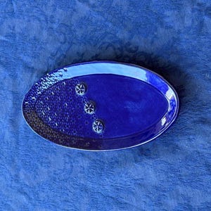 May include: An oval ceramic dish in a deep cobalt blue glaze. The surface features a smooth, glossy finish on the right side and a textured, dimpled pattern on the left. Three small, raised flower-shaped accents are aligned in the center.