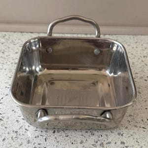 May include: A square, polished stainless steel serving dish featuring rounded corners and two arched metal handles attached to opposite sides. The interior base is flat, and the walls are vertical, showcasing a reflective, metallic silver finish.