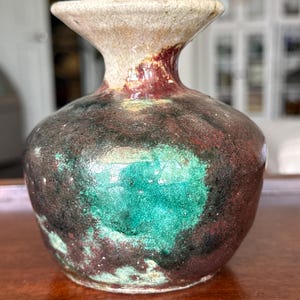 Può includere: Piccolo vaso in ceramica fatto a mano con forma organica. Presenta una tavolozza di colori terrosi macchiati, con bordo color crema, collo bordeaux scuro e corpo marmorizzato marrone scuro e verde acqua. Base bulbosa e apertura svasata.