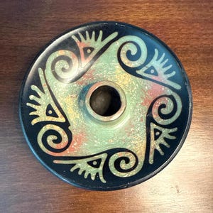 Puede incluir: Un portavelas redondo de cerámica negra con un agujero central. El portavelas presenta un centro moteado verde, amarillo y rojo y está decorado con diseños negros repetidos alrededor del borde.