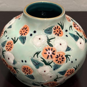 May include: A round, light green ceramic vase with a floral design. The vase features orange and white flowers with dark green leaves. The rim is gold, and the interior is dark green. A decorative piece for the home.
