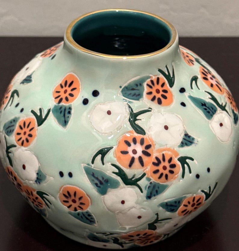 May include: A round, light green ceramic vase with a floral design. The vase features orange and white flowers with dark green leaves. The rim is gold, and the interior is dark green. A decorative piece for the home.