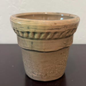 Puede incluir: Una pequeña maceta de cerámica de estilo antiguo en color verde-marrón apagado. La maceta tiene una superficie texturizada y una banda decorativa en forma de cuerda alrededor de la parte superior. La maceta está sobre una superficie oscura.