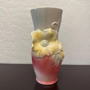 May include: A ceramic vase with a floral design. The vase has a light gray top, transitioning to a pink base. A large yellow flower with green leaves is sculpted on the front. The vase is approximately 7 inches tall.