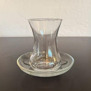 May include: A clear glass tea cup with a distinct tulip-shaped silhouette, featuring a narrow waist and a flared rim, resting on a matching circular clear glass saucer. The set is made of transparent, smooth glass with no patterns or decorations.