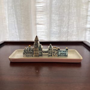 May include: A ceramic trinket tray featuring a miniature London skyline. The tray is rectangular with a light pink border. The skyline includes iconic landmarks in black and white with gold accents.