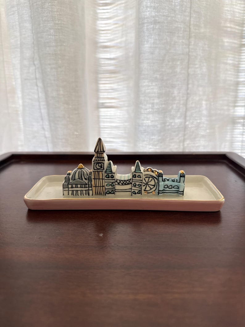 May include: A ceramic trinket tray featuring a miniature London skyline. The tray is rectangular with a light pink border. The skyline includes iconic landmarks in black and white with gold accents.