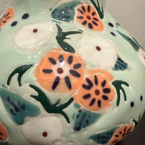 May include: A ceramic vase with a light green base color. The vase is decorated with orange and white floral designs, green leaves, and dark blue accents. The vase has a glossy finish and a rounded shape.