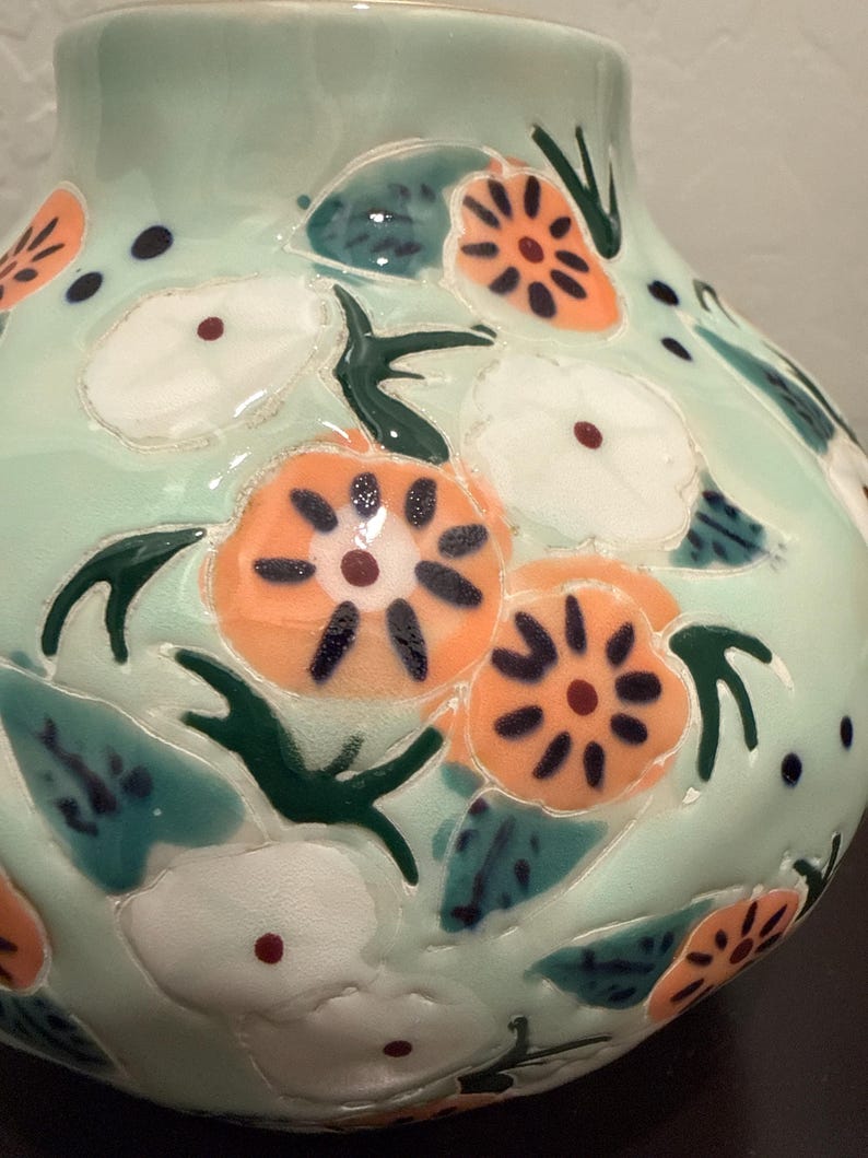 May include: A ceramic vase with a light green base color. The vase is decorated with orange and white floral designs, green leaves, and dark blue accents. The vase has a glossy finish and a rounded shape.