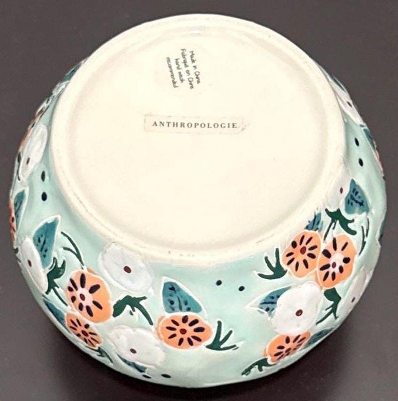May include: A round, light blue ceramic bowl with a floral design. The bowl features white and orange flowers with green leaves. The bottom of the bowl is white and has the word "ANTHROPOLOGIE" printed on it.