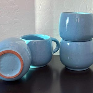 May include: Four blue ceramic mugs with rounded bodies and handles. Two mugs are stacked, and the others are arranged nearby. The interior of the mugs is a lighter blue. The bottom of one mug shows a reddish-brown ring.