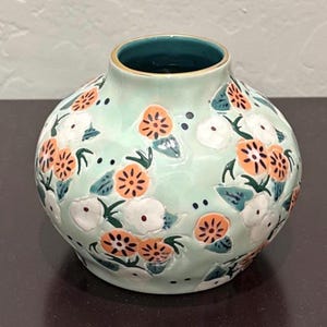 May include: A round, ceramic vase with a light green base, adorned with orange and white floral patterns and dark green leaves. The vase has a dark teal interior and a gold rim.