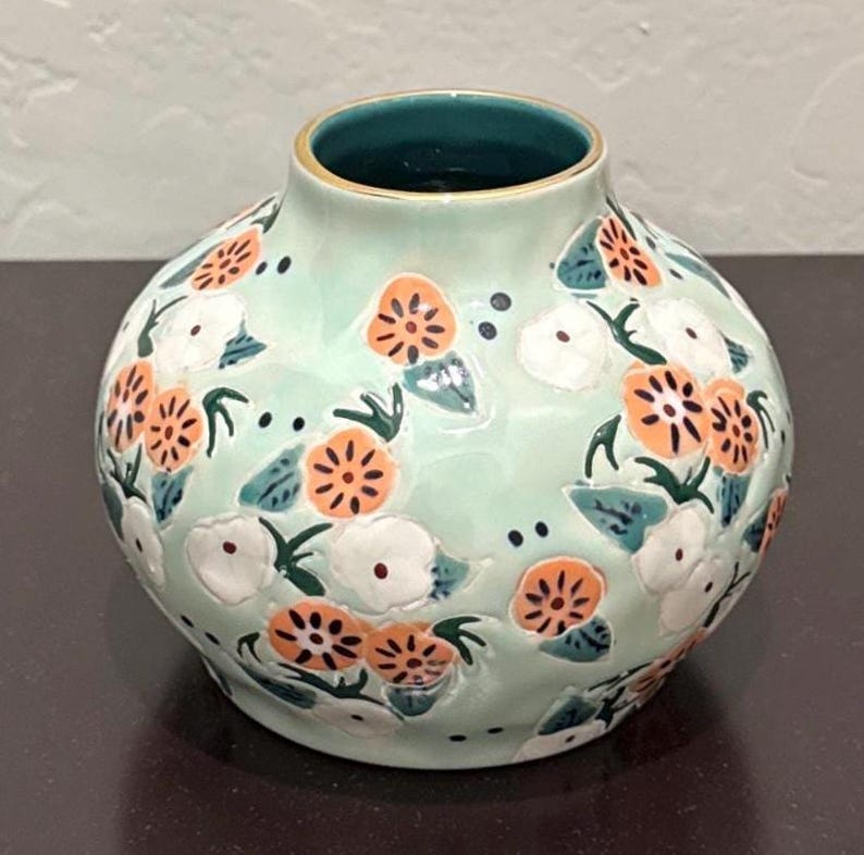May include: A round, ceramic vase with a light green base, adorned with orange and white floral patterns and dark green leaves. The vase has a dark teal interior and a gold rim.