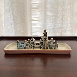May include: A ceramic trinket dish with a pink rim and a London skyline design. The skyline includes the London Eye, Big Ben, and other iconic buildings. The dish is rectangular and sits on a dark wooden surface.