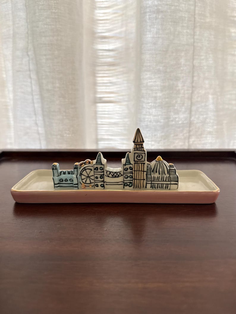 May include: A ceramic trinket dish with a pink rim and a London skyline design. The skyline includes the London Eye, Big Ben, and other iconic buildings. The dish is rectangular and sits on a dark wooden surface.