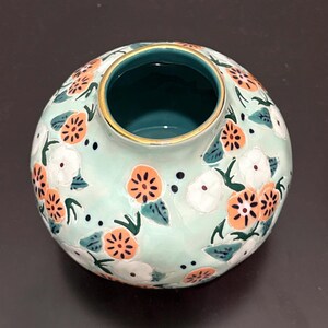 May include: A round, ceramic vase with a light teal background and a floral pattern. The flowers are orange and white with green leaves. The rim is gold, and the inside is teal. The vase is on a dark surface.