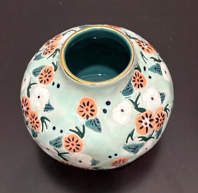 May include: A round, ceramic vase with a light teal background and a floral pattern. The flowers are orange and white with green leaves. The rim is gold, and the inside is teal. The vase is on a dark surface.