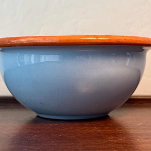 May include: A light blue ceramic bowl with a rounded shape and a contrasting orange rim. The bowl is sitting on a wooden surface. The bowl is likely used for serving food.