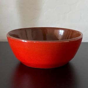 May include: A small, round ceramic bowl with a vibrant red exterior and a dark brown interior. The bowl is set against a neutral background, highlighting its simple, yet striking design. This bowl is suitable for serving small portions of food.
