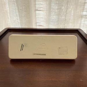 May include: A rectangular, off-white box with the word "London" printed in a stylized font. The word "ANTHROPOLOGIE" is printed below. The box is on a dark brown wooden surface.