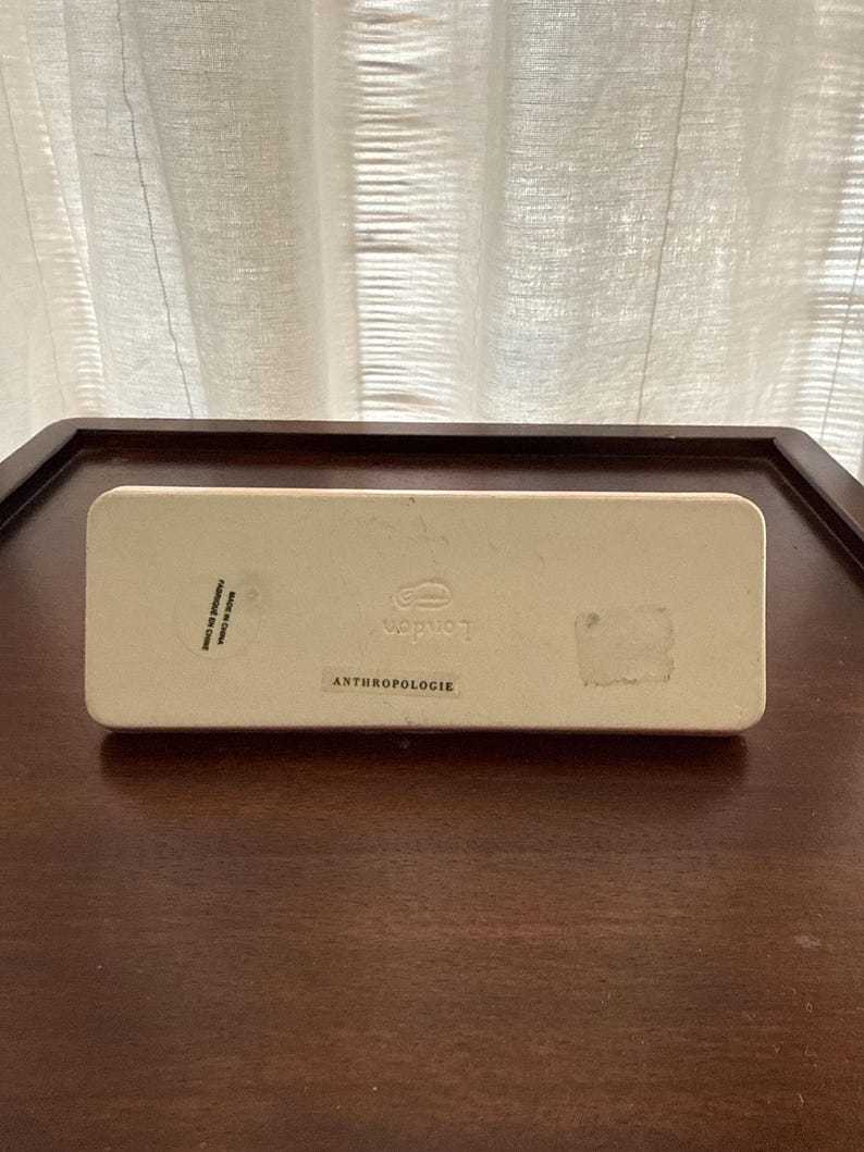 May include: A rectangular, off-white box with the word "London" printed in a stylized font. The word "ANTHROPOLOGIE" is printed below. The box is on a dark brown wooden surface.