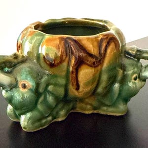Puede incluir: Una maceta de cerámica con forma de elefante, con un esmalte verde, marrón y amarillo. La maceta tiene una abertura ancha y dos asas con forma de trompas de elefante. La maceta mide aproximadamente 15 cm de largo, 10 cm de ancho y 10 cm de alto.