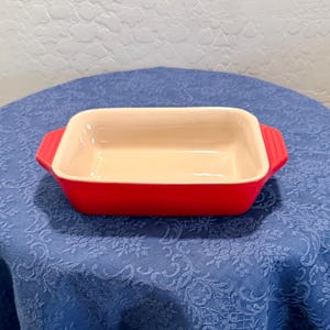Op de afbeelding: Een rechthoekige ovenschaal met een felrode buitenkant en een gladde, crèmekleurige binnenkant. De schaal is voorzien van twee geïntegreerde handgrepen aan de zijkanten met een geribbelde textuur voor een stevige grip bij het verplaatsen.
