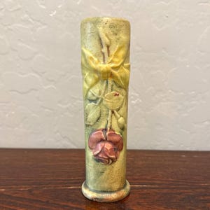 May include: A cylindrical ceramic vase with a matte finish in shades of muted green and yellow. The exterior features a raised relief design of a single red rose blossom, green leaves, and a yellow ribbon tied in a bow near the top rim.