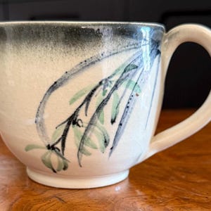 May include: Cream-colored ceramic mug with a rounded body and a large handle. The mug features a hand-painted design of black and green floral branches. The top rim is painted in a dark gray color.
