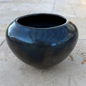 May include: A round, dark blue ceramic bowl with a glossy finish. The bowl has a wide opening and a smooth, curved body. The surface reflects light, creating highlights and shadows. The bowl is likely used for decorative purposes.