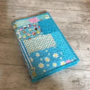 May include: A rectangular fabric book cover featuring a patchwork design in shades of blue, white, and pink. The quilted surface displays various patterns including cupcakes, white daisies, blue gingham checks, and polka dots, finished with a blue border.