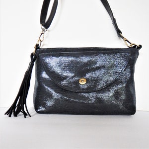 Może przedstawiać: Czarna skórzana torebka na ramię z srebrnym połyskiem i frędzlem. Torebka ma klapkę zamykaną złotym guzikiem.