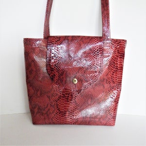 Może przedstawiać: Czerwona torba typu tote ze skóry wężowej z klapką i jednym zatrzaskiem. Torba ma dwa paski na ramię.