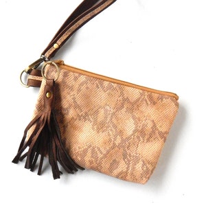 Może przedstawiać: Mała, beżowa skórzana torebka z brązowym skórzanym paskiem i frędzlem. Torebka ma wzór skóry węża i zamek błyskawiczny.