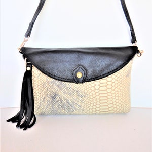 Könnte beinhalten: Eine schwarze und weiße Leder-Umhängetasche mit Schlangenhautmuster. Die Tasche hat einen Klappenverschluss mit einem goldenen Knopf und einem schwarzen Quasten.