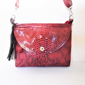 Może przedstawiać: Czerwona torebka na ramię ze skóry węża z klapką i czarną frędzlą. Torebka ma złote zapięcie na guzik.