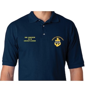 May include: A navy blue polo shirt with a yellow embroidered emblem featuring an anchor and the text "WORLD'S GREATEST NAVY USN". The shirt also has the text "USS HANCOCK CV-19 AIRCRAFT CARRIER" embroidered on the left chest.