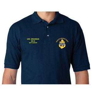 May include: A navy blue polo shirt with the text "USS ARKANSAS BB-33 BATTLESHIP" embroidered on the left chest and "WORLD'S GREATEST NAVY USN" embroidered on the right chest.