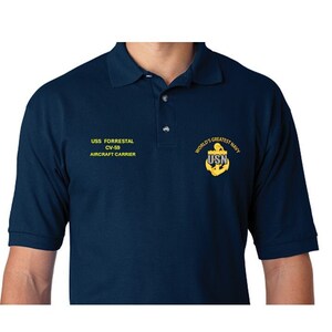 Könnte beinhalten: Ein marineblaues Poloshirt mit gelbem gesticktem Text, der "USS FORRESTAL CV-59 AIRCRAFT CARRIER" lautet, und einem gelben gestickten Anker mit dem Text "WORLD'S GREATEST NAVY USN".